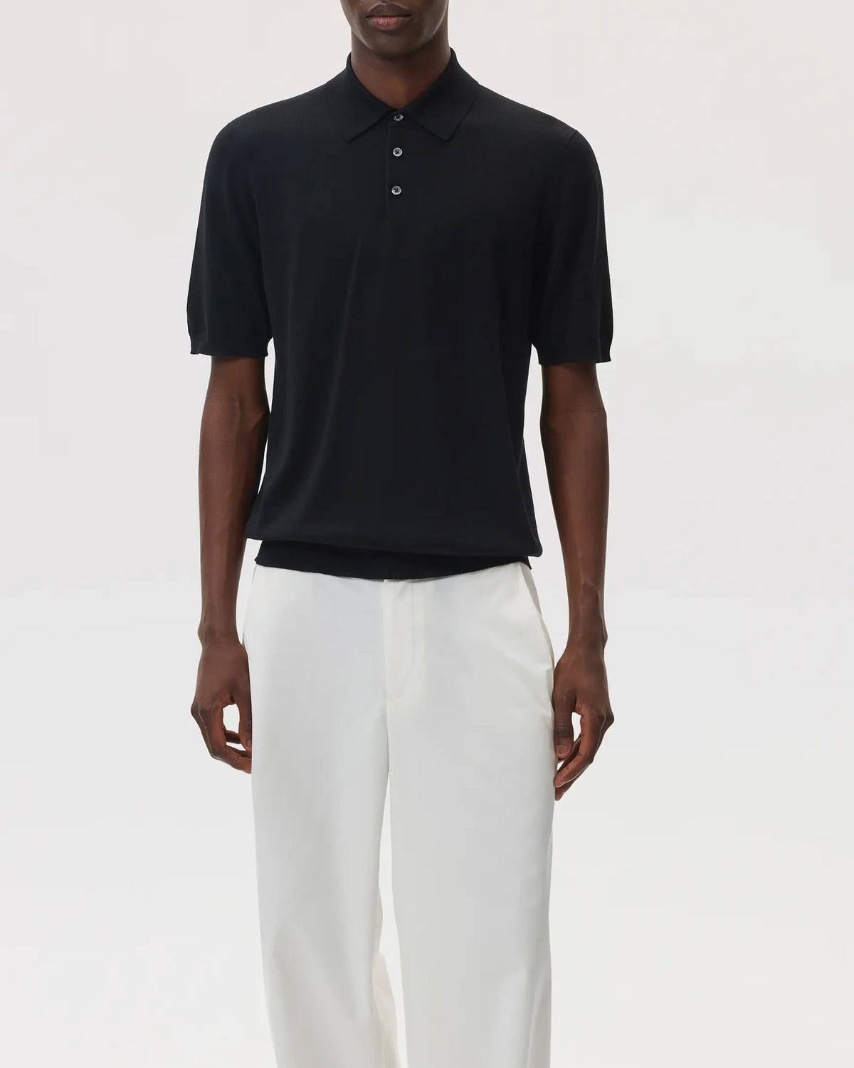 Model wears Johnstons of Elgin SS25 Men's Knitwear Black Superfine Merino Polo Shirt with white trousers on a pale grey background KDI00715SA7131