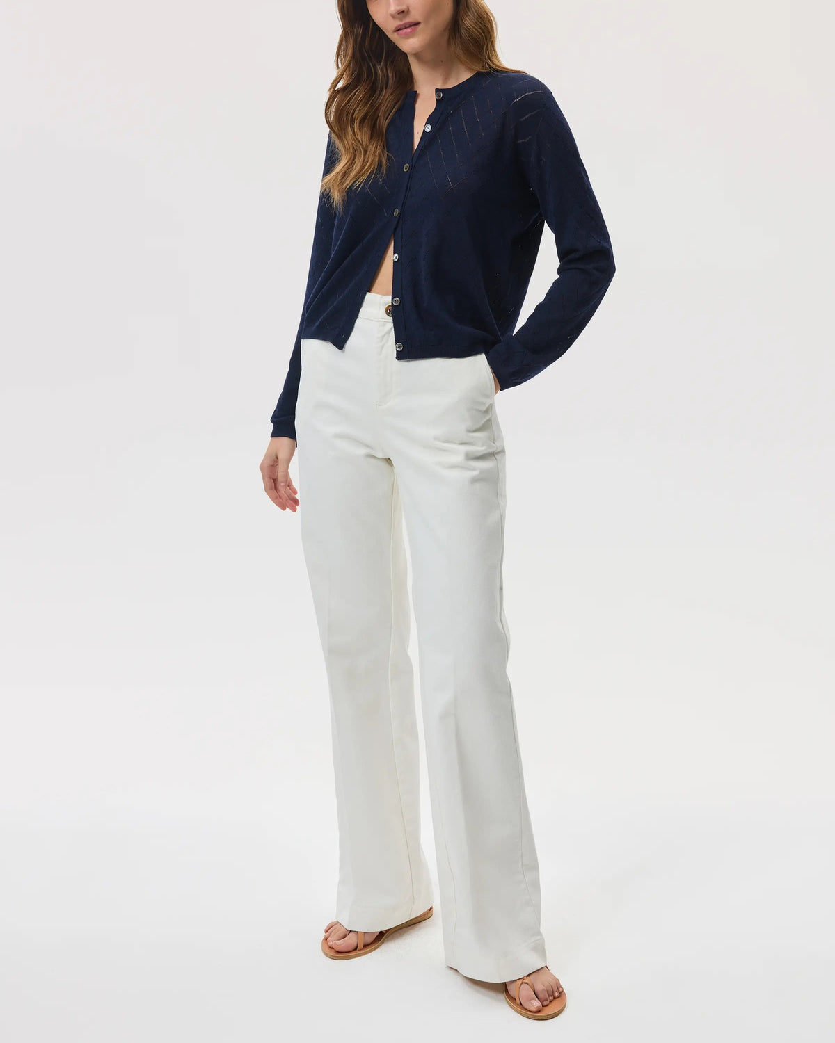 Model wears Johnstons of Elgin SS25 Women's Knitwear Navy Superfine Merino Pointelle Argyle Cardigan with white trousers and sandals KDI00742SD7563