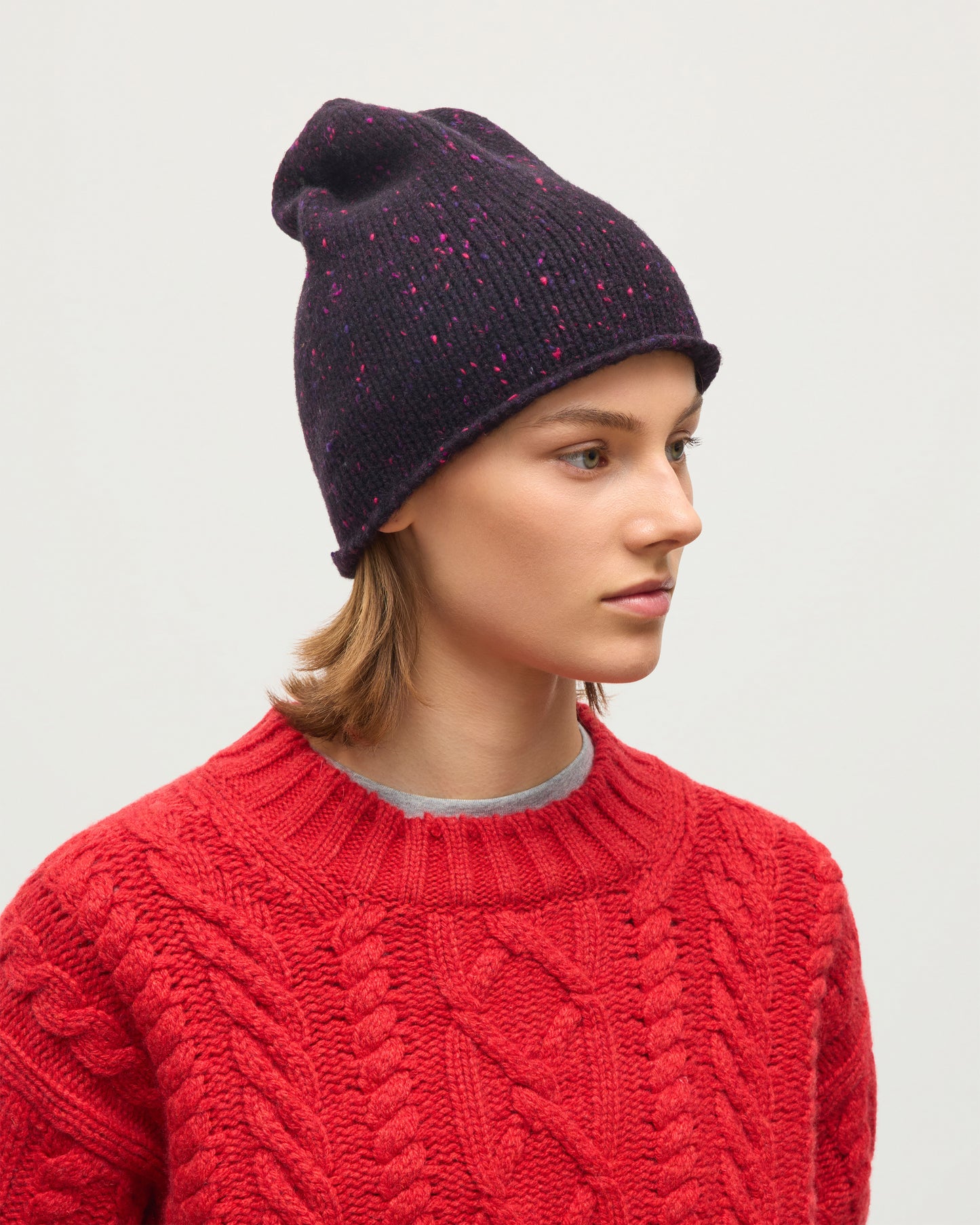 A woman in profile wearing a dark plum knit beanie with pink speckles and a red cable-knit sweater, photographed against a light gray studio background. JOHNSTONS OF ELGIN CASHMERE DONEGAL TIREE BEANIE PURPLE 100% CASHMERE HAC03613HD7360ONE
