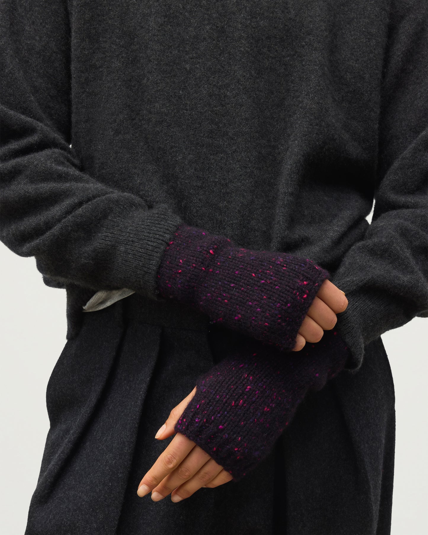 Close-up of a person wearing dark charcoal knit sleeves with fingerless cuffs in a deep purple tone with small pink flecks, shown against a plain light background. JOHNSTONS OF ELGIN DONEGAL CASHMERE TIREE WRISTWARMERS PURPLE 100% CASHMERE HAC03644HD7360ONE