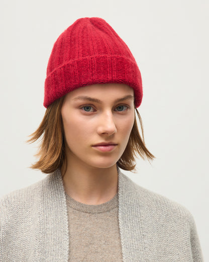 A woman with short brown hair wearing a bright red ribbed knit beanie and a light gray cardigan over a beige sweater, photographed against a plain light gray background. JOHNSTONS OF ELGIN BRUSHED CASHMERE SILK FLAT RIB BHALTAIR BEANIE RED 74% CASHMERE, 26% SILK HBC01196HE4389ONE