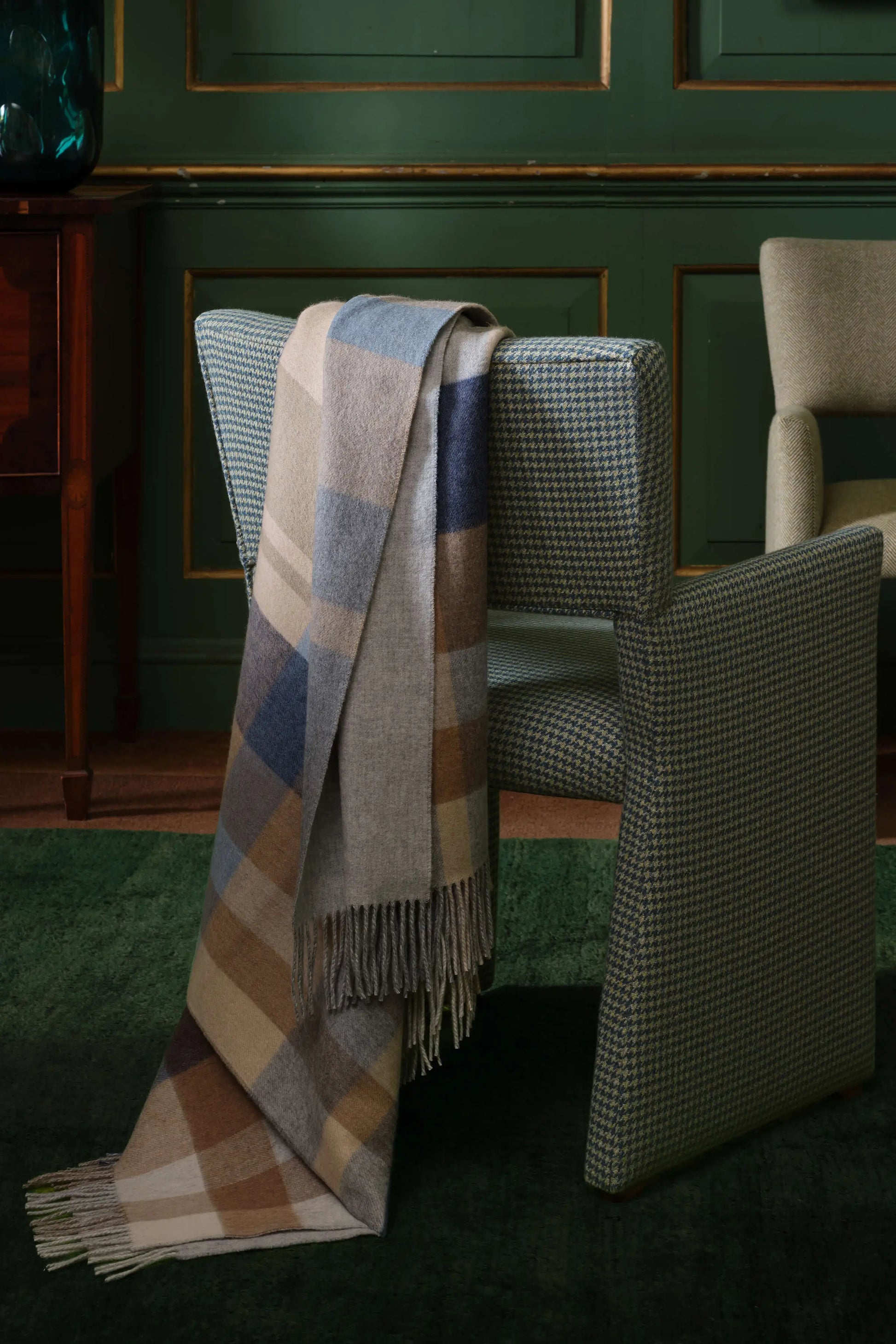 A stylish chair featuring a Johnstons of Elgin reversible cashmere throw in Birch Check, positioned in front of a lush green wall, creating a warm atmosphere. WA000013RU7603ONE