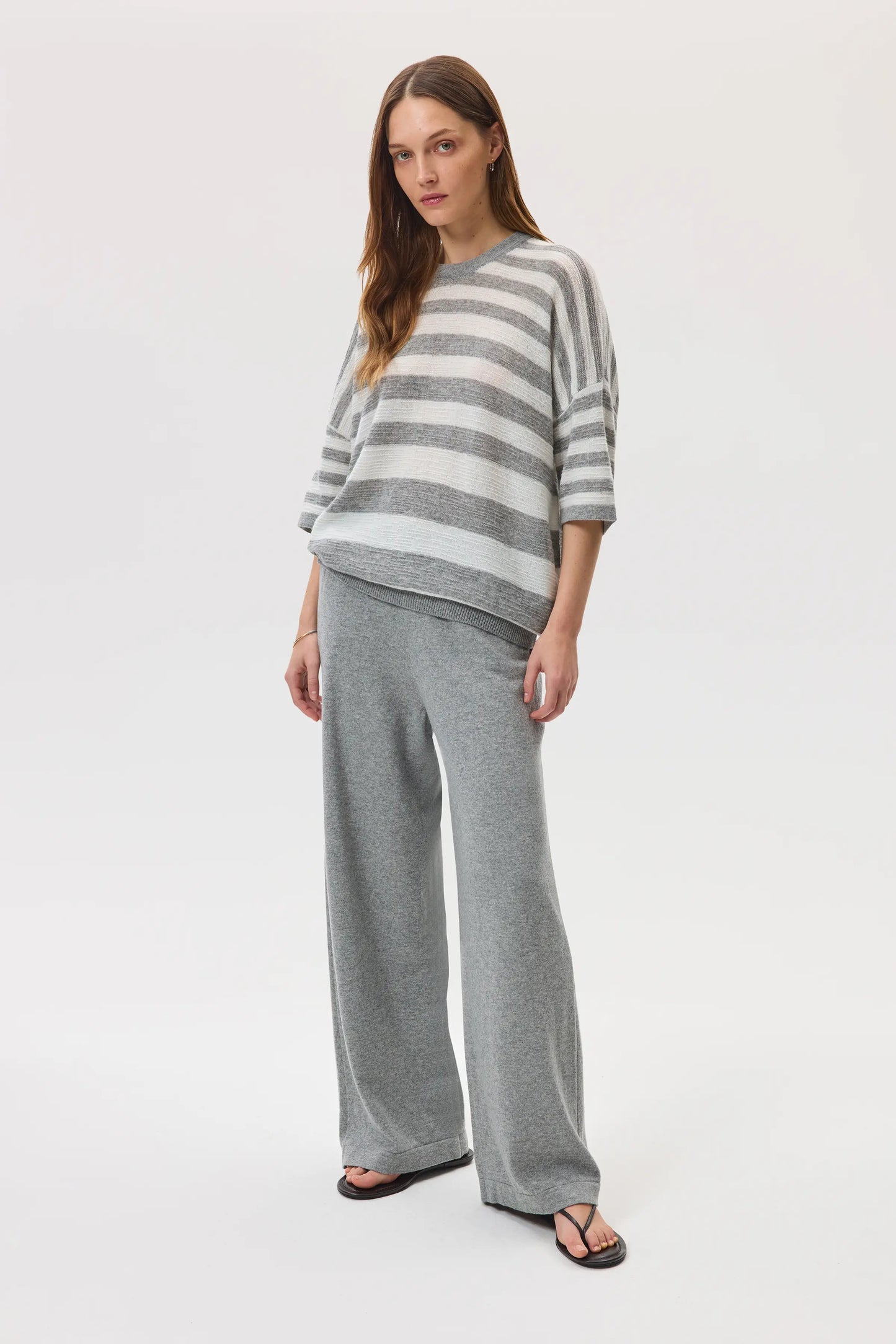Female model wearing Johnstons of Elgin SS25 Unisex Knitwear Light Grey & White Textured Stripe Oversized Cashmere T-Shirt with grey wide leg trousers KAA05414Q25124