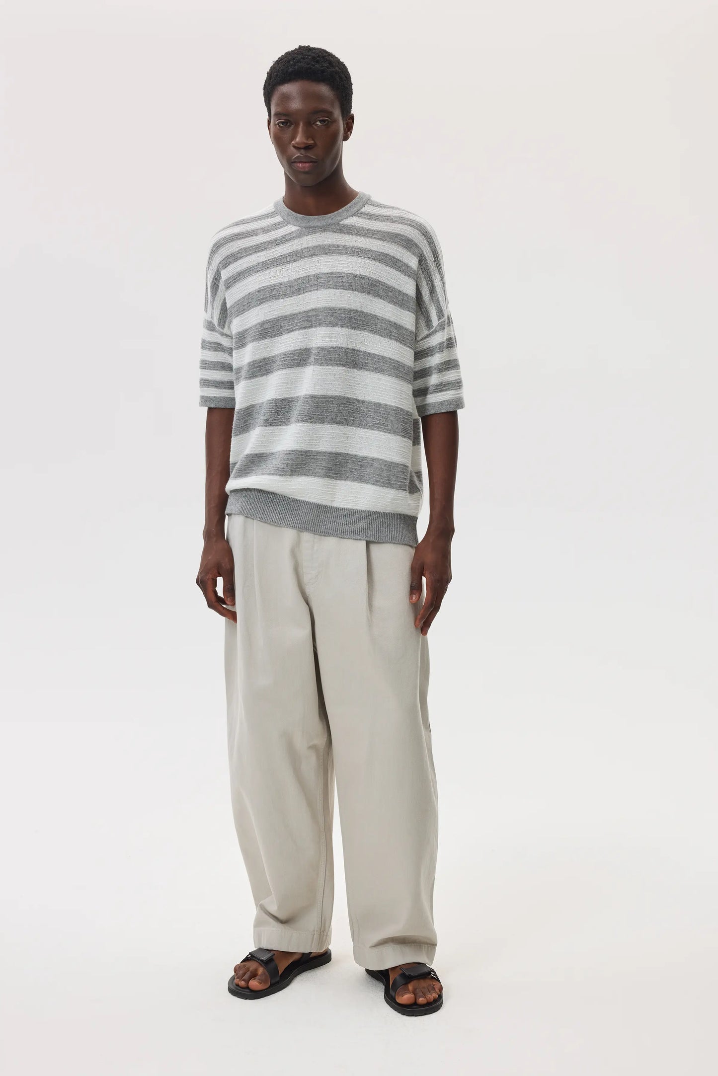 Male model wearing Johnstons of Elgin SS25 Unisex Knitwear Light Grey & White Textured Stripe Oversized Cashmere T-Shirt with cream trousers and sandals on a white backdrop KAA05414Q25124