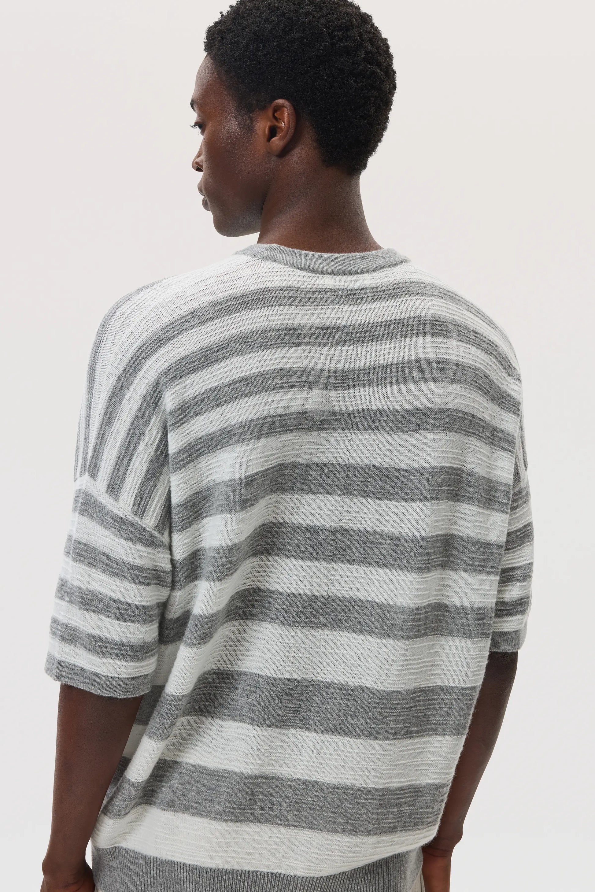 Back view of a male model wearing Johnstons of Elgin SS25 Unisex Knitwear Light Grey & White Textured Stripe Oversized Cashmere T-Shirt on a white backdrop KAA05414Q25124