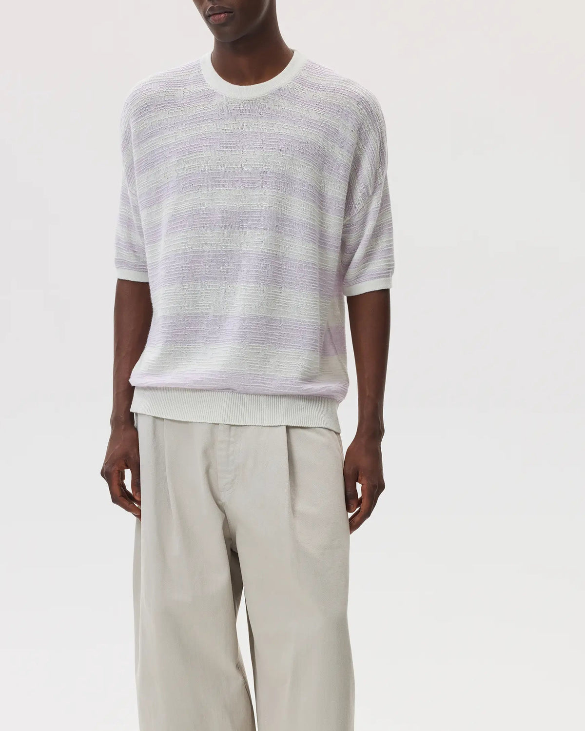 Male model wearing Johnstons of Elgin SS25 Unisex Knitwear White & Lavender Textured Stripe Oversized Cashmere T-Shirt with cream trousers and black sandals on a white background KAA05414Q25126