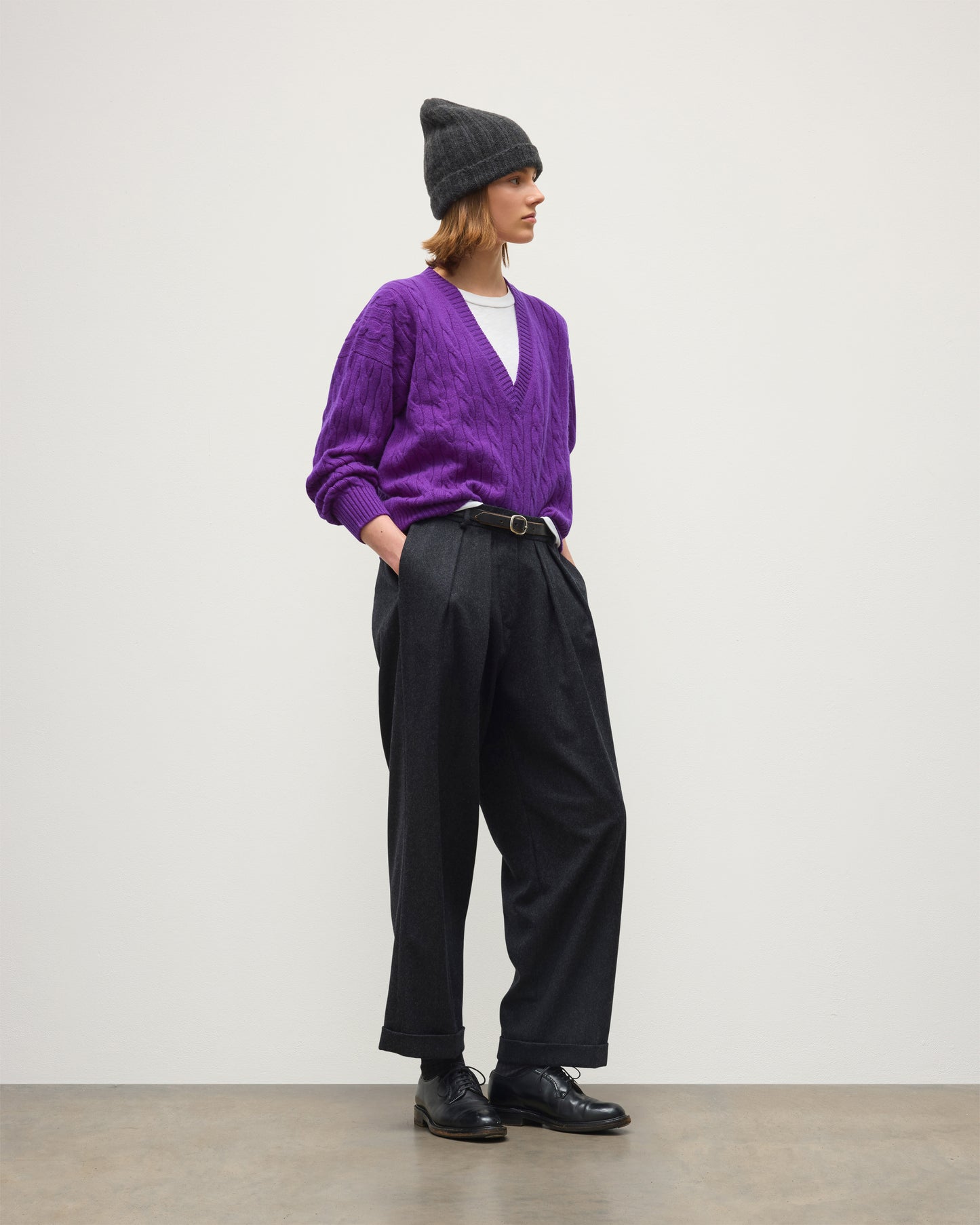 A model stands in profile wearing a purple cable-knit V-neck sweater, black wide-leg trousers, black leather shoes, a black beanie, and a thin black belt with a gold buckle, set against a minimalist off-white studio background. JOHNSTONS OF ELGIN KYLESKU DEEP VEE SWEATER CASHMERE CABLE DEEP VEE SWEATER PURPLE 100% CASHMERE KAA05728SD5372