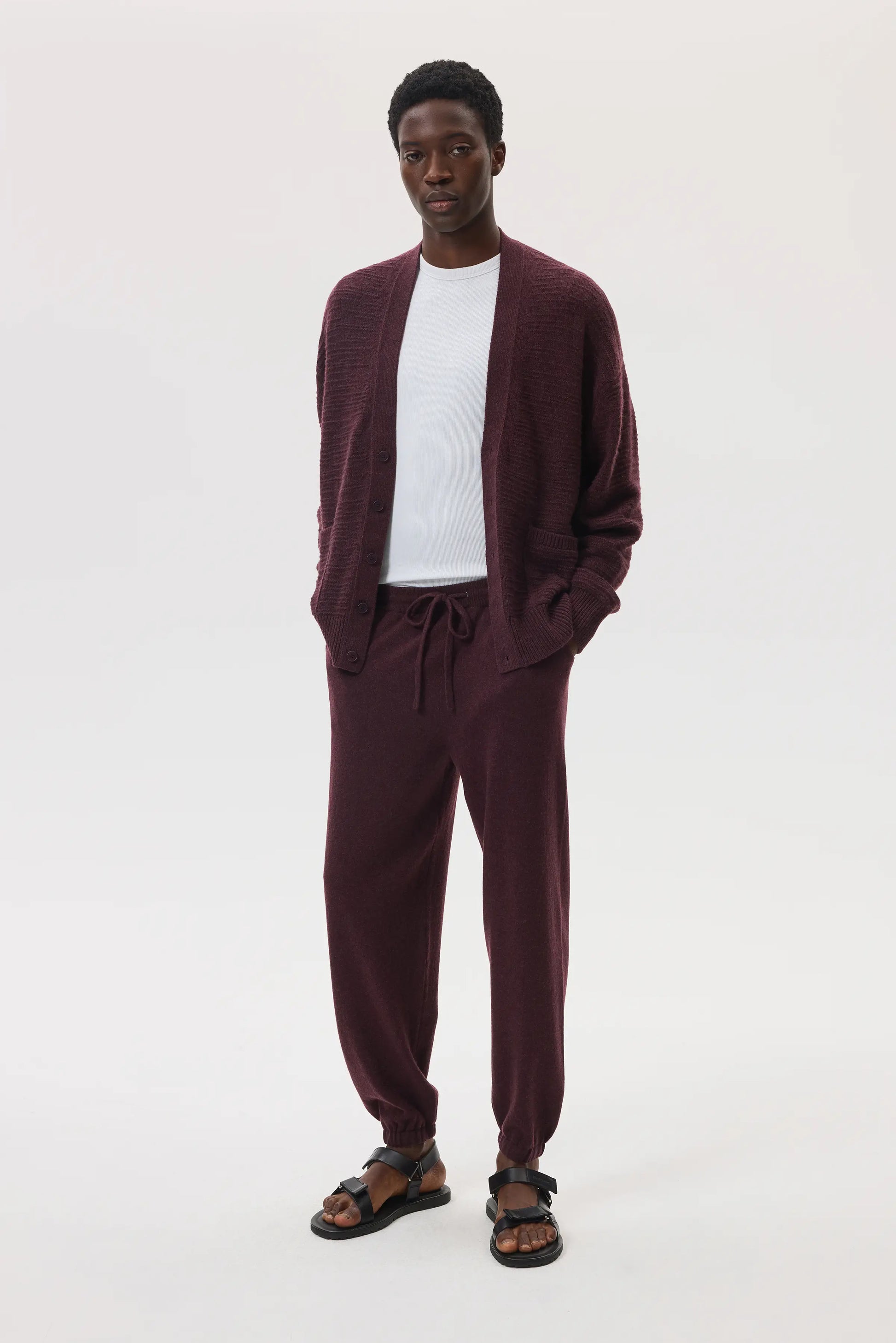 Male model wearing Johnstons of Elgin SS25 Unisex Knitwear Rosewood Textured Cashmere Cardigan with superfine cashmere sweater in white, and rosewood cashmere jogger against a white background KAC05415SB7089