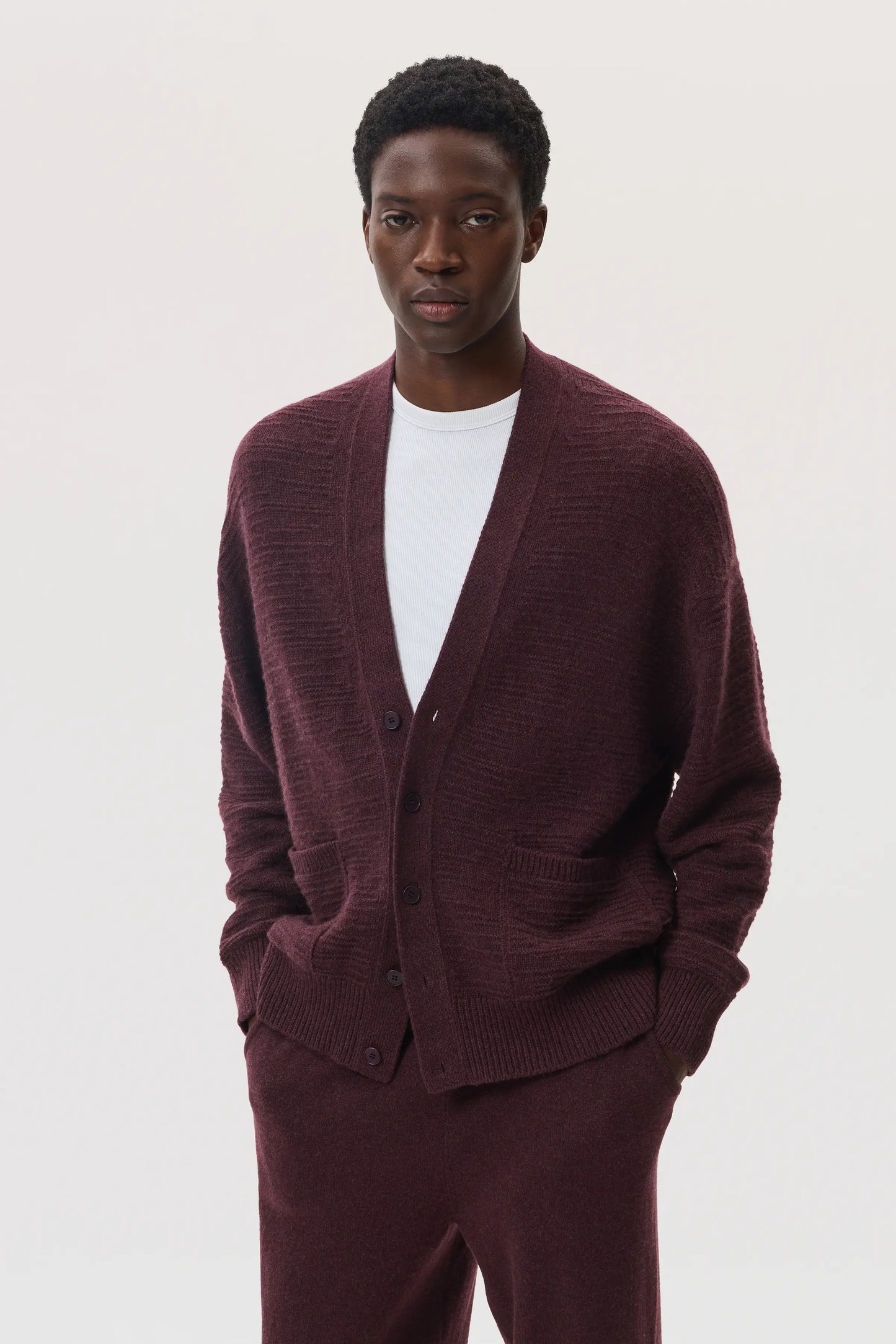 Male model wearing Johnstons of Elgin SS25 Unisex Knitwear Rosewood Textured Cashmere Cardigan with superfine cashmere sweater in white, and rosewood cashmere jogger  KAC05415SB7089