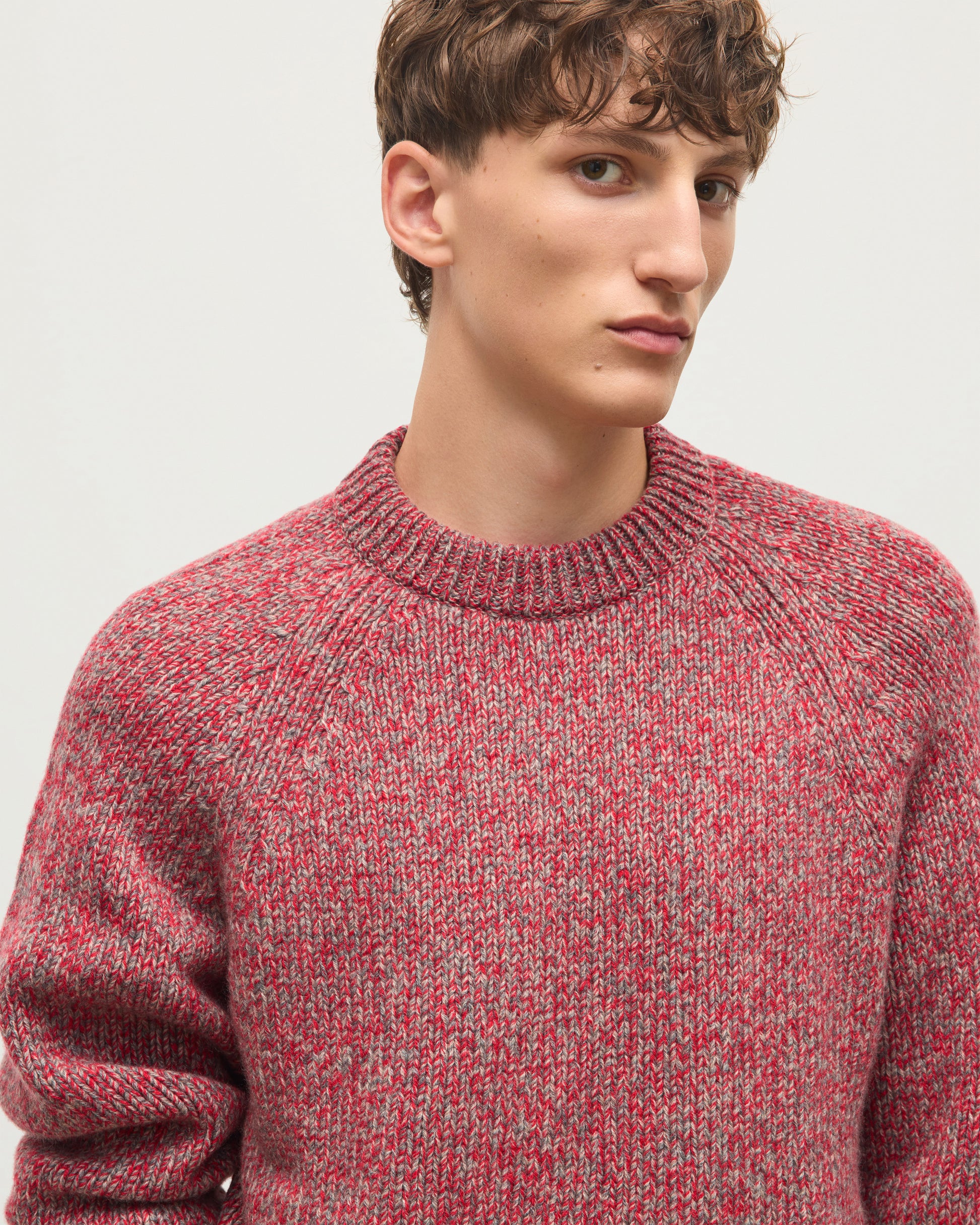 A close-up of a young man wearing a textured, heathered knit crewneck sweater in red and gray, photographed against a plain light gray background. JOHNSTONS OF ELGIN FYFE CREW SWEATER CASHMERE 3 COLOUR MARL CREW SWEATER - 6 PLY RED MARL 100% CASHMERE KAC05753007014