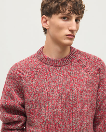 A close-up of a young man wearing a textured, heathered knit crewneck sweater in red and gray, photographed against a plain light gray background. JOHNSTONS OF ELGIN FYFE CREW SWEATER CASHMERE 3 COLOUR MARL CREW SWEATER - 6 PLY RED MARL 100% CASHMERE KAC05753007014