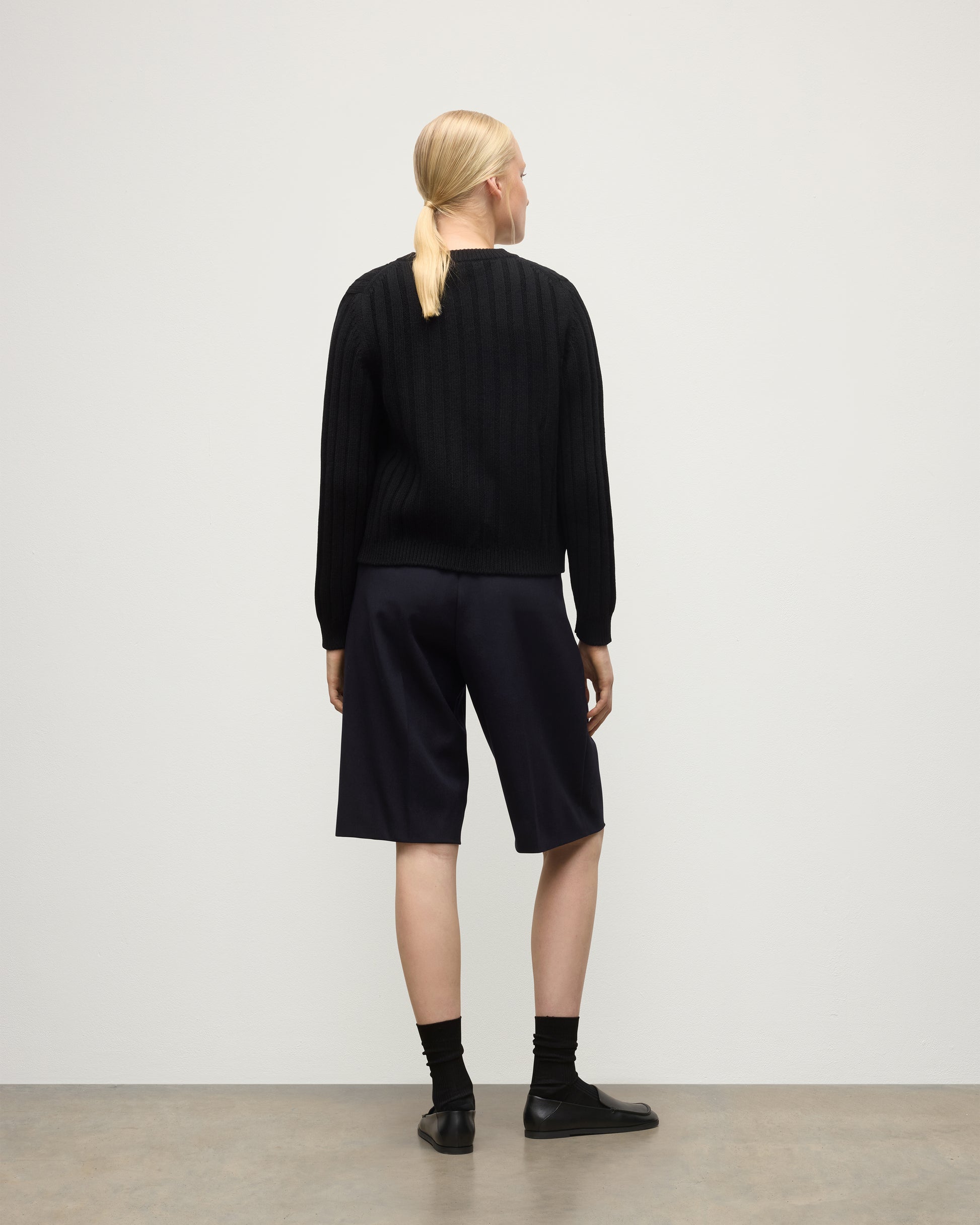 BACK OF MODEL WEAR JOHNSTONS OF ELGIN WOMENS LECHT CREW CARDIGAN BLACK 100% CASHMERE OVER A WHITE T-SHIRT & LONG NAVY SHORTS KAC05790SA7210