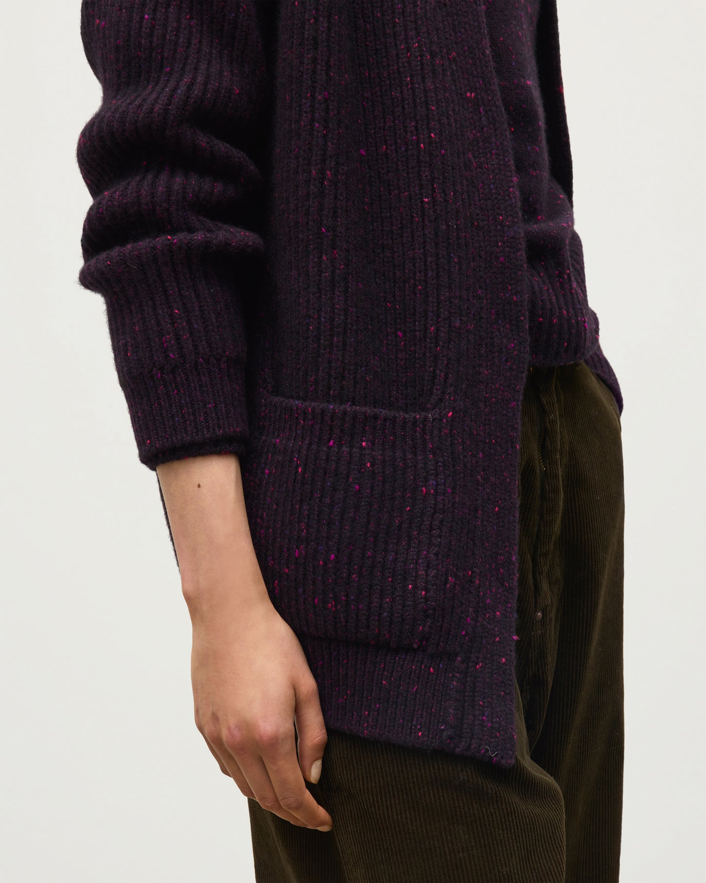 Close-up of a person wearing a dark purple ribbed knit sweater with subtle pink and blue flecks, paired with dark brown corduroy pants against a plain light background. JOHNSTONS OF ELGIN NORAH CARDIGAN DONEGAL CASHMERE RIBBED CARDIGAN WITH POCKETS DARK PURPLE 100% CASHMERE KAC05992HD7360