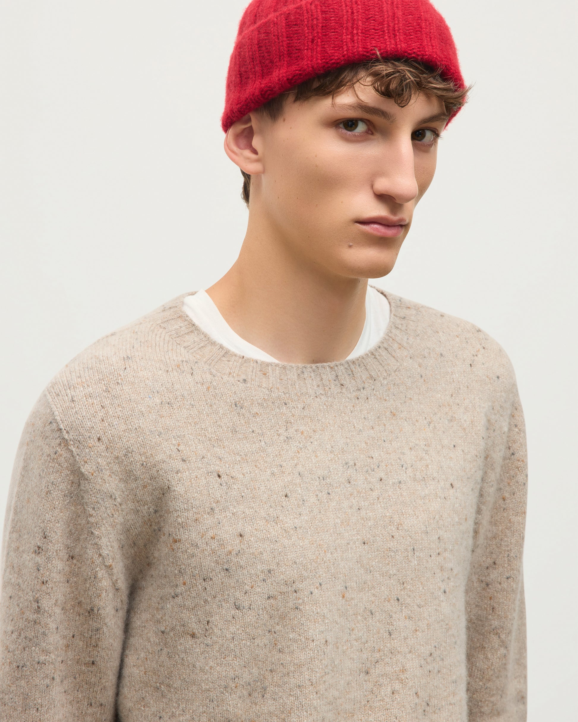 A man wearing a red knit beanie and a light beige speckled crewneck sweater over a white undershirt, photographed against a plain light gray background. JOHNSTONS OF ELGIN TIREE CREW SWEATER DONEGAL CASHMERE CREW SWEATER CREAM 100% CASHMERE KAI05755HA0397