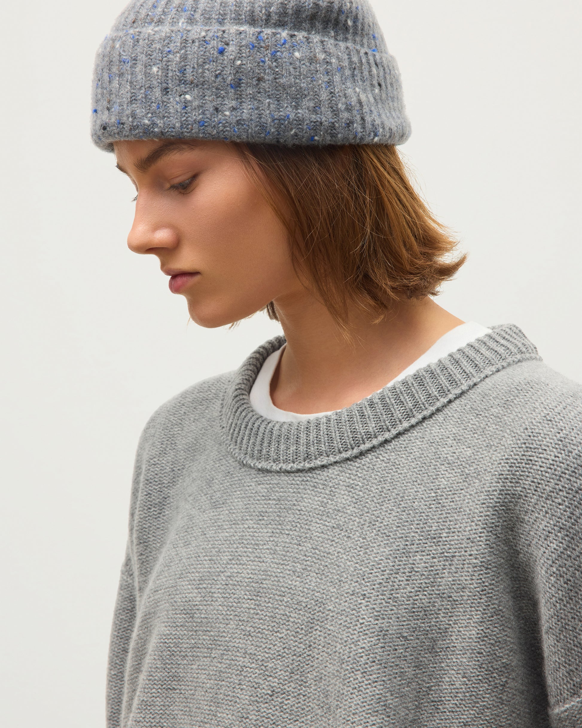A close-up of a person wearing a gray knit crewneck sweater and a speckled gray beanie against a plain light background. JOHNSTONS OF ELGIN MAGGIE CREW SWEATER CASHMERE REVERSE STITCH ROUND NECK SWEATER - 8 PLY GREY 100% CASHMERE KAI05990HA0400