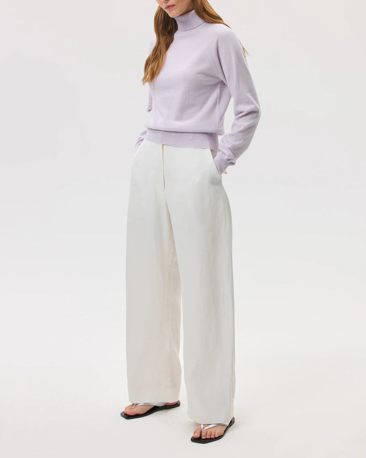 Model wears Johnstons of Elgin SS25 Women's Knitwear Lavender Classic Cashmere Roll Neck Jumper with white trousers on a white backdrop KAP05275HD0086