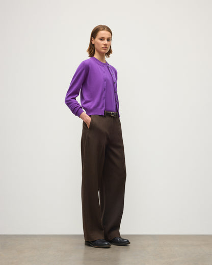 A model stands in a studio wearing a purple long-sleeve button-front cardigan, dark brown wide-leg trousers, a black belt, and black shoes against a light gray background. JOHNSTONS OF ELGIN IRIS CREW CARDIGAN PETITE CASHMERE ROUND NECK CARDIGAN - 2 PLY PURPLE 100% CASHMERE KAP05704SD5371