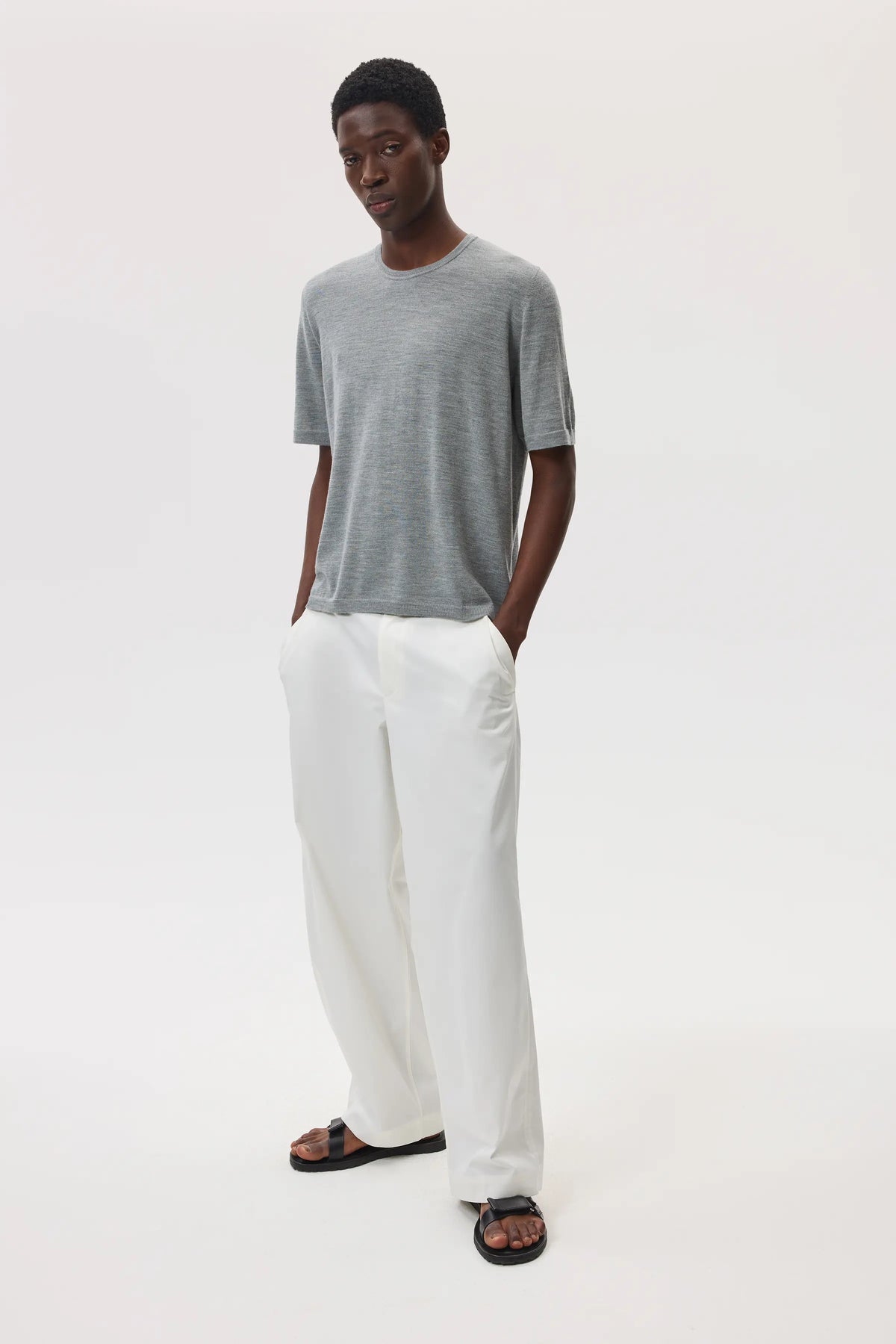 Model wears Johnstons of Elgin SS25 Men's Knitwear Light Grey Superfine Merino T-Shirt with white trousers against a white studio backdrop KDI00663HA4183