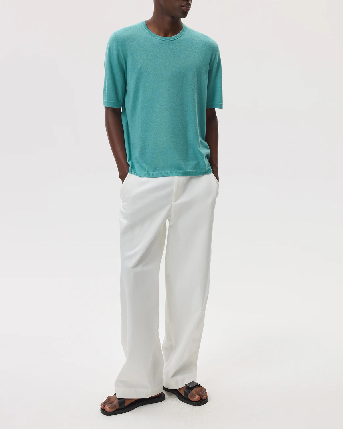 Model wearing Johnstons of Elgin SS25 Men's Knitwear Jade Superfine Merino T-Shirt with white trousers against a white backdrop KDI00663SC4641